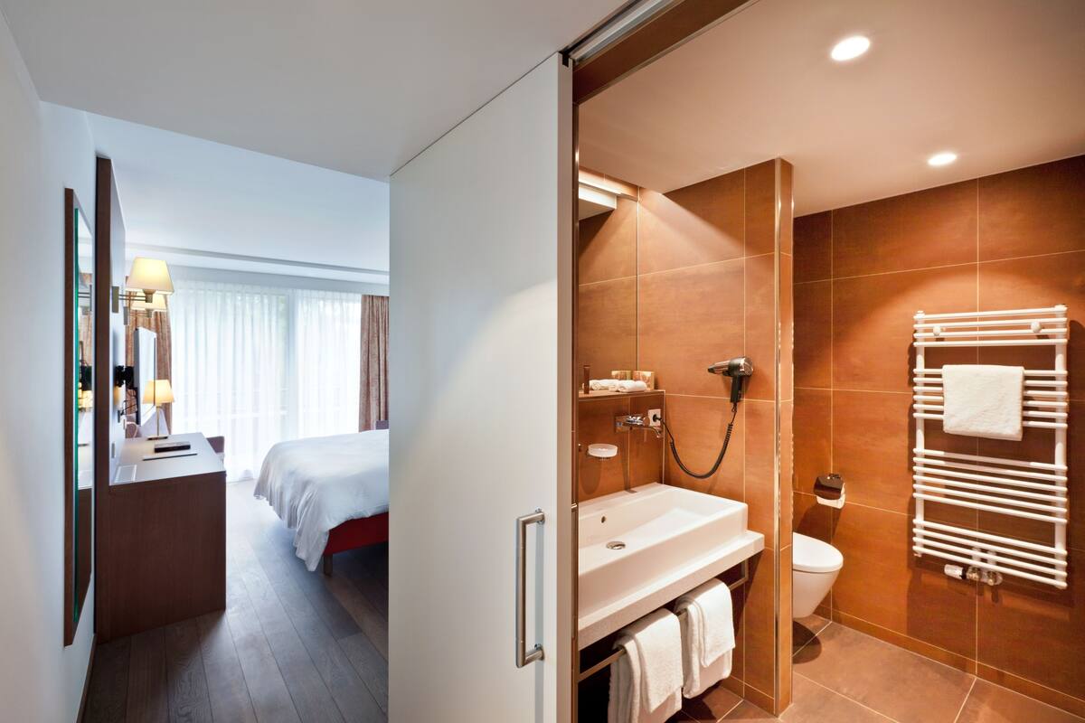 Una habitación de hotel moderna con una cama, un baño con lavamanos, inodoro y perchero para toallas.