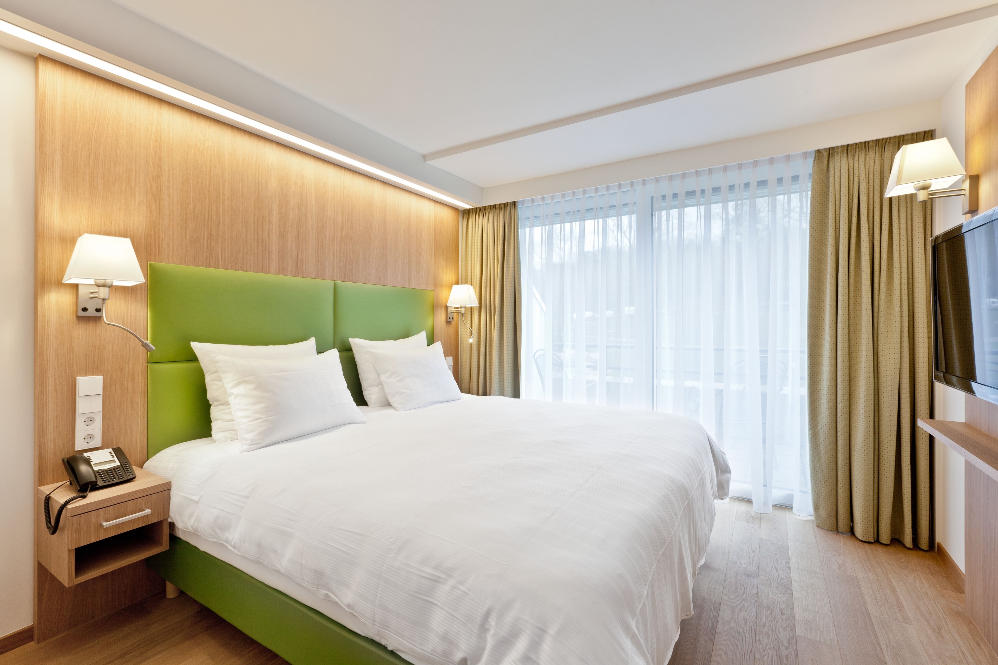 Quarto de hotel com cabeceira verde, roupa de cama branca, telefone ao lado da cama e televisão de tela plana sobre um suporte de madeira.