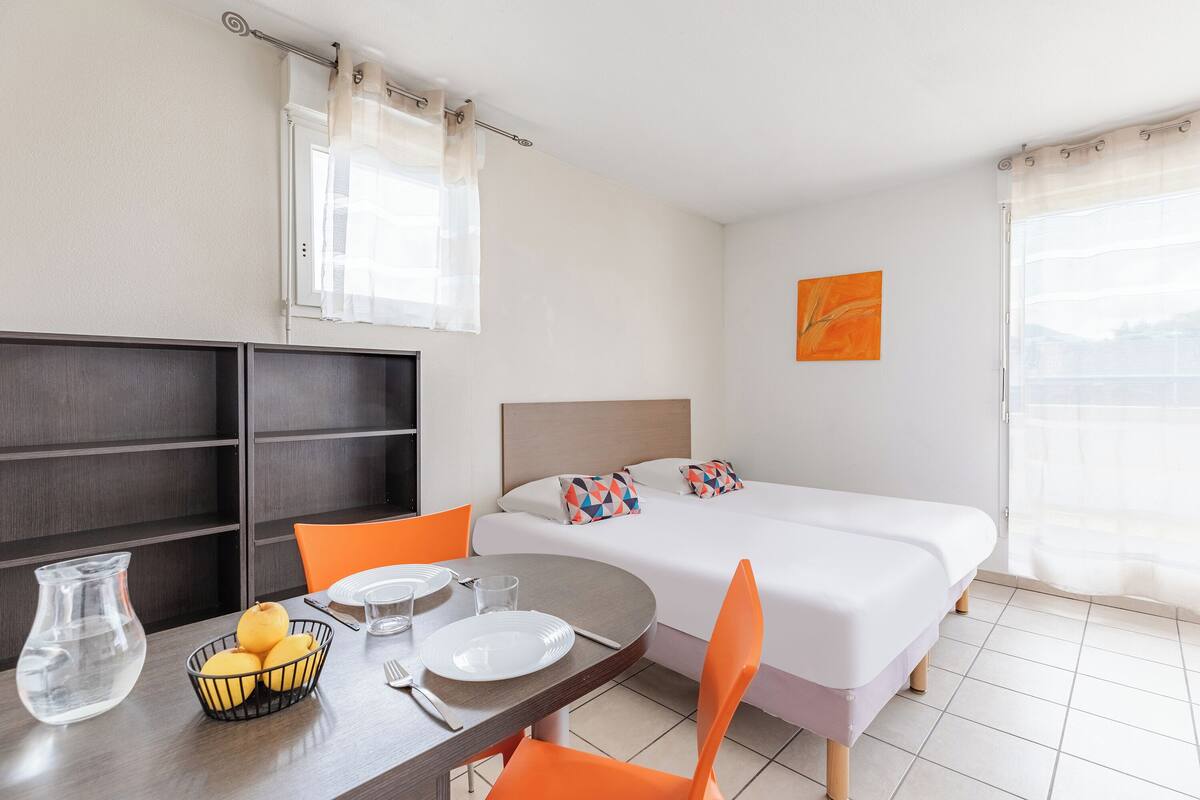 Une chambre d’hôtel comprenant un lit, une table à manger avec des chaises, un petit coin cuisine et une fenêtre avec des rideaux.