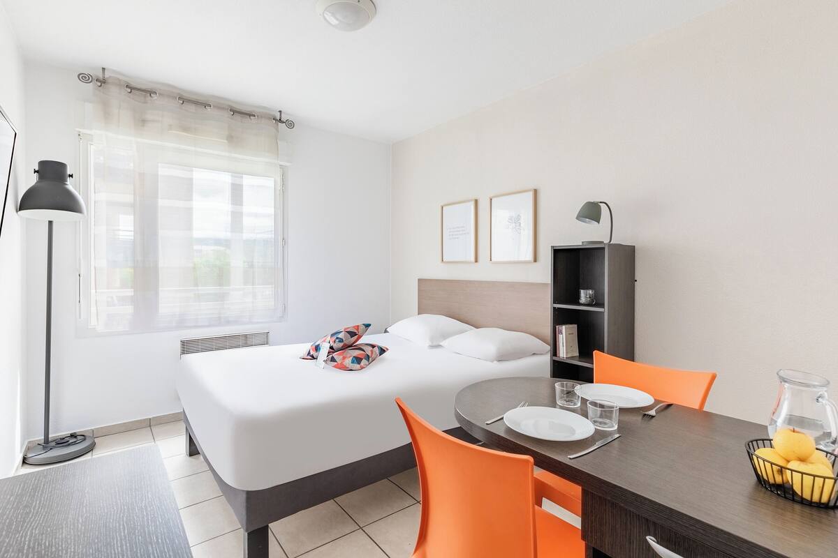 Une chambre d’hôtel comprenant un lit, une table à manger avec des chaises, une bibliothèque et une fenêtre avec des rideaux.