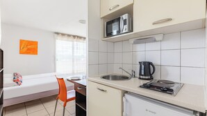 A compact kitchen with a microwave, sink, and small refrigerator. There is a window with curtains, a sofa with a colorful pillow, and a chair. - Appart'City Classic Thonon les bains (Thonon-les-Bains)