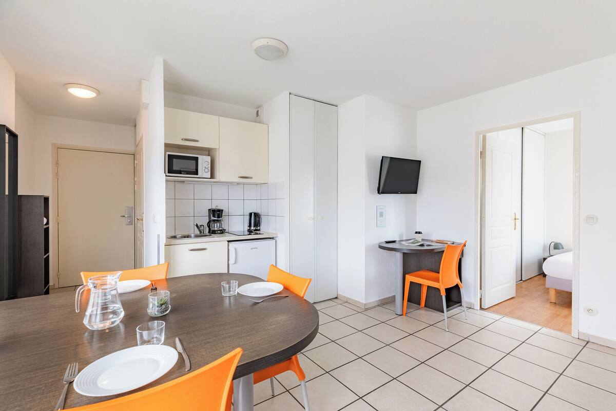Une cuisine moderne avec des meubles blancs, un four à micro-ondes et une machine à café. On y trouve une table à manger avec des chaises orange et un téléviseur fixé au mur.