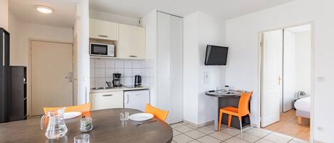 Una cocina moderna con armarios blancos, un microondas y una cafetera. Hay una mesa de comedor con sillas anaranjadas y un televisor montado en la pared.