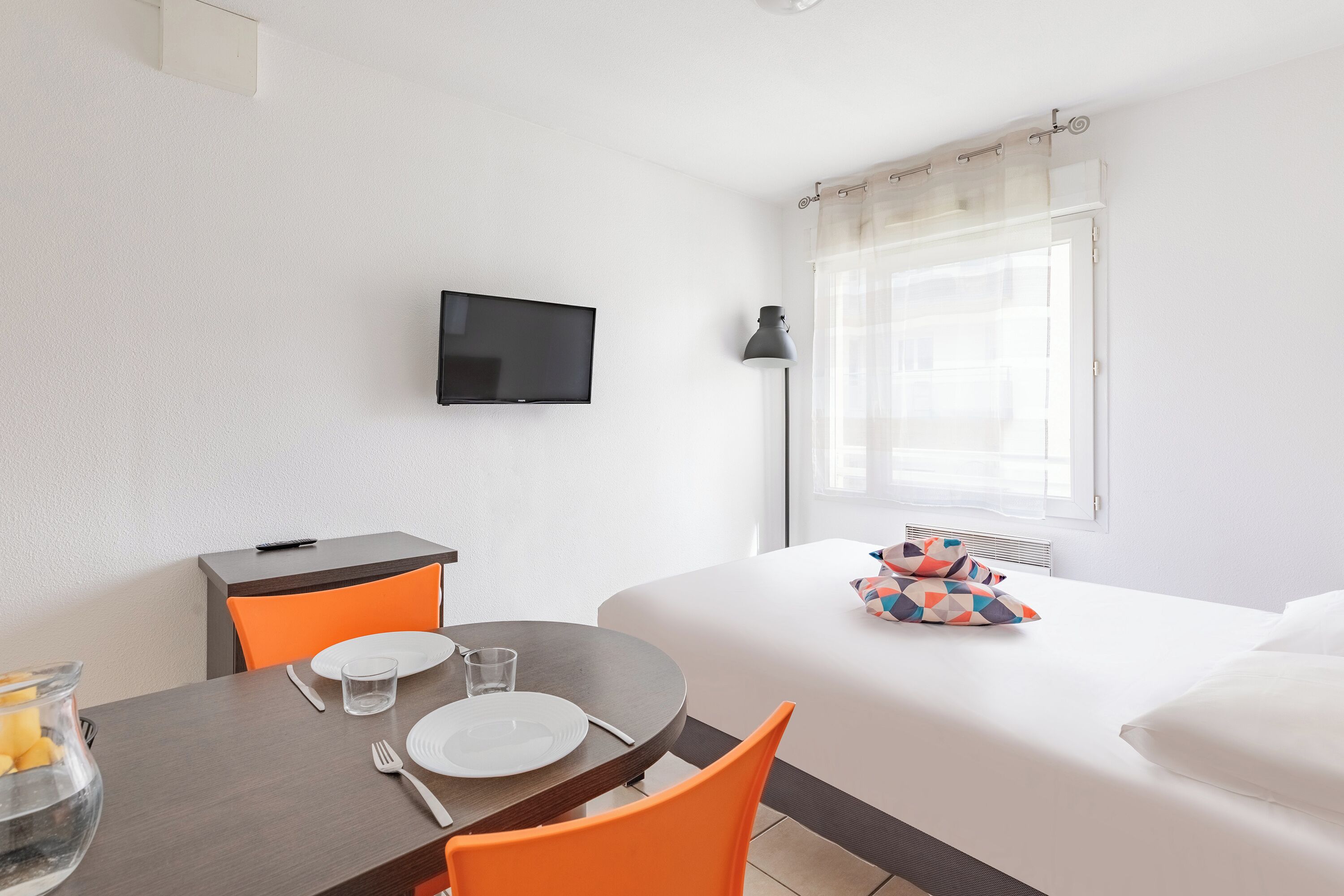 Ein Hotelzimmer mit Bett, Wand-Fernseher, Esstisch mit Stühlen und einem Vorhangfenster.