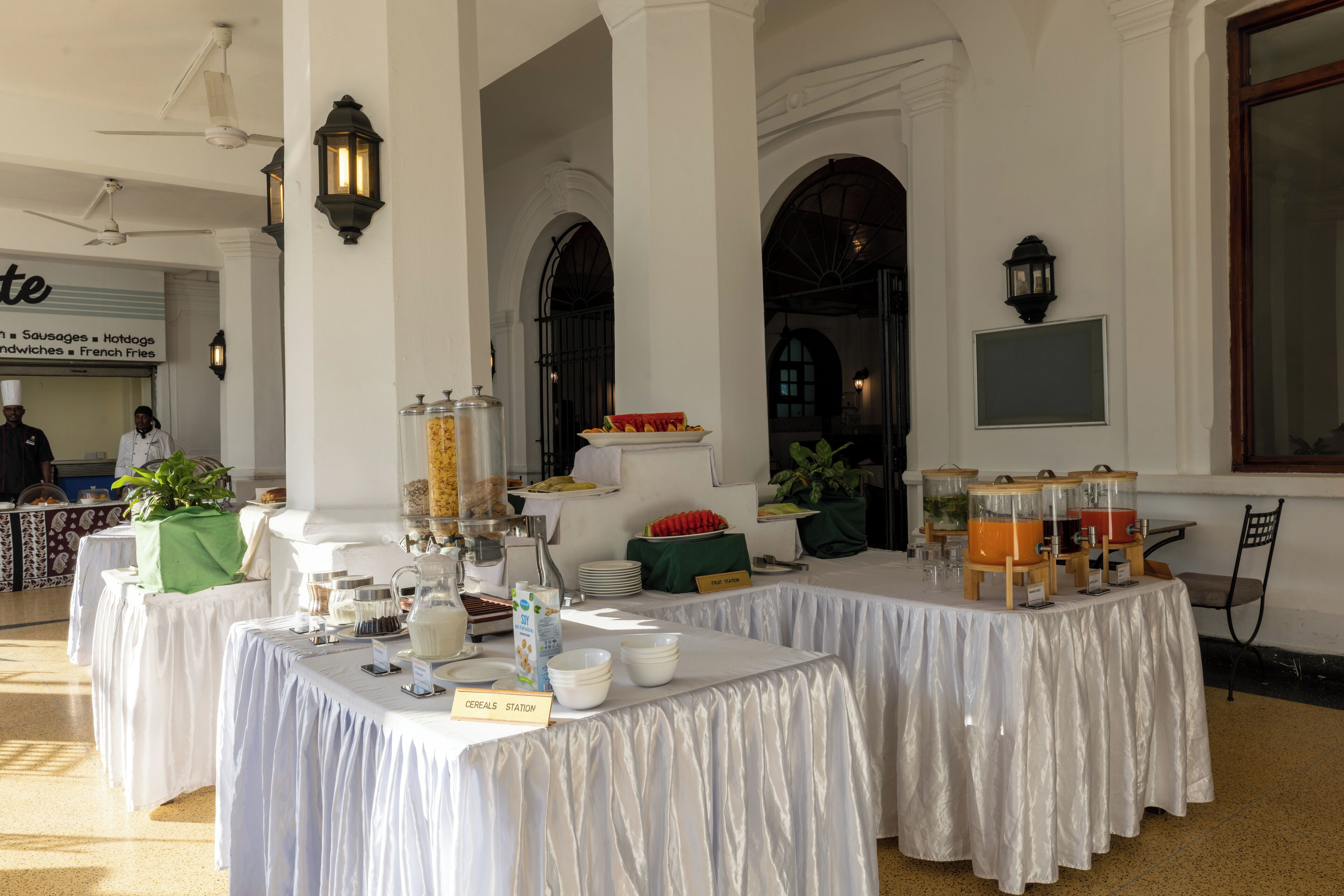 Un buffet de petit-déjeuner avec différents stands de nourriture, notamment un stand de céréales, et un espace repas avec des tables et des chaises.