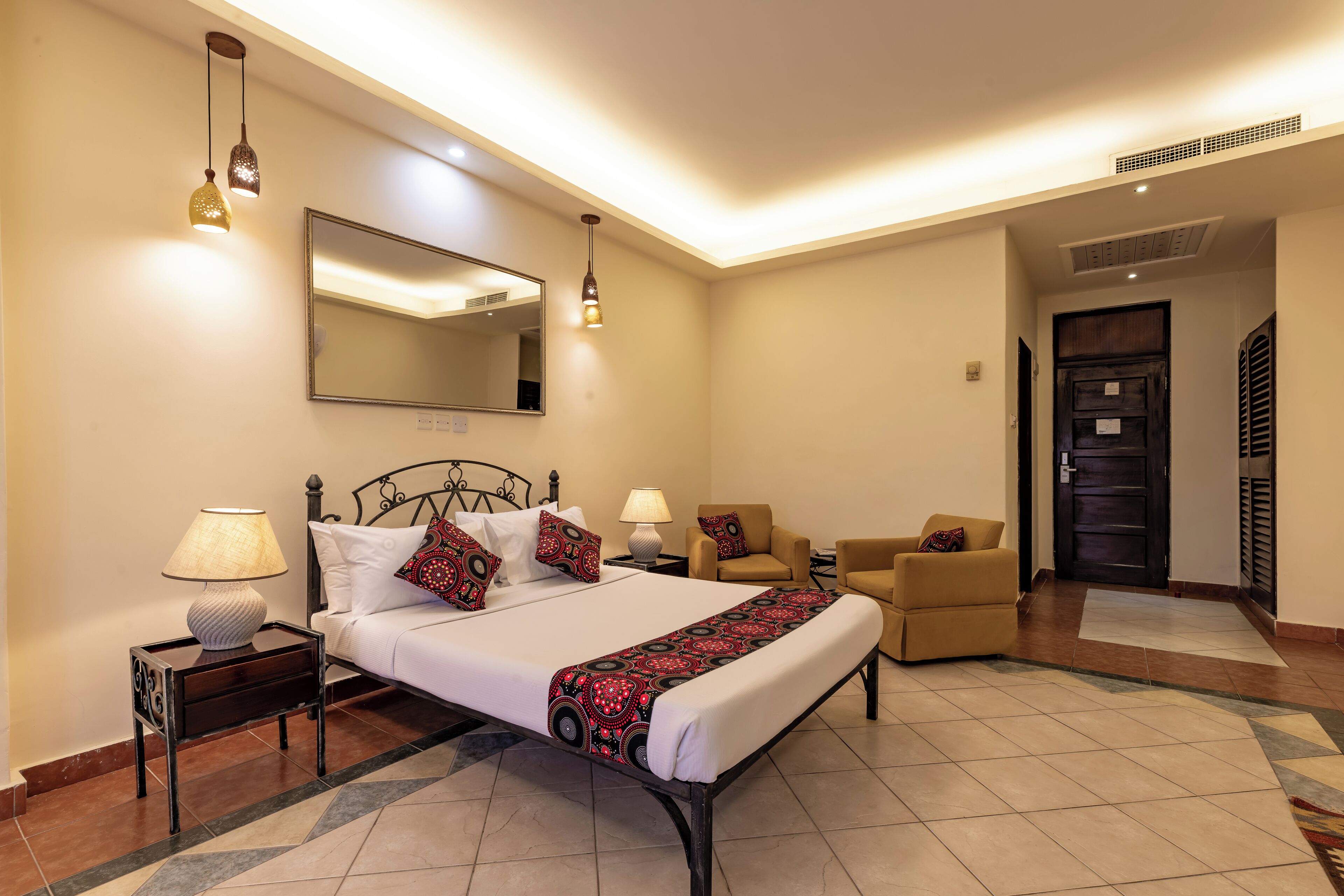 A hotel room with a bed, two chairs, a nightstand, a lamp, a mirror, and a door.