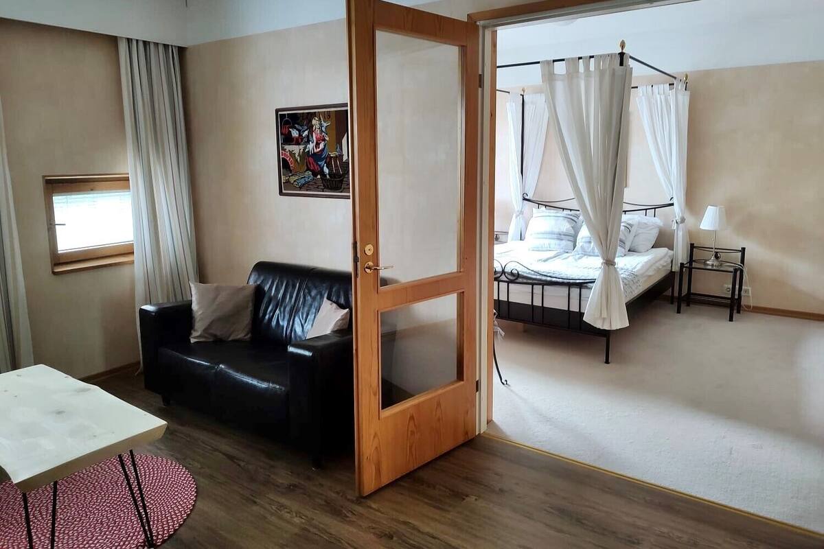 Una habitación con sofá de cuero negro, una mesita, una cama con dosel y una ventana con cortinas.