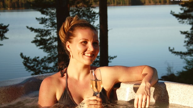 Une personne dans un bain à remous, un verre de champagne à la main, entourée d’arbres et d’une étendue d’eau.
