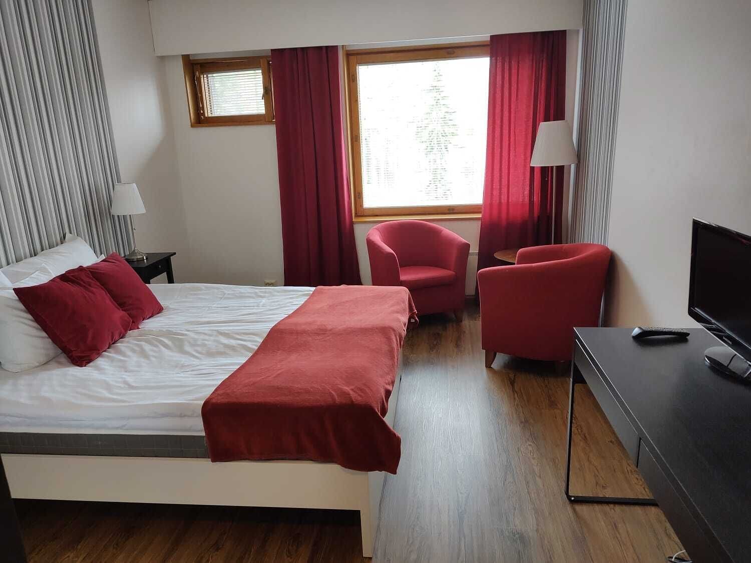 Habitación de hotel con cama, dos sillas rojas, escritorio y televisión, y ventana con cortinas.
