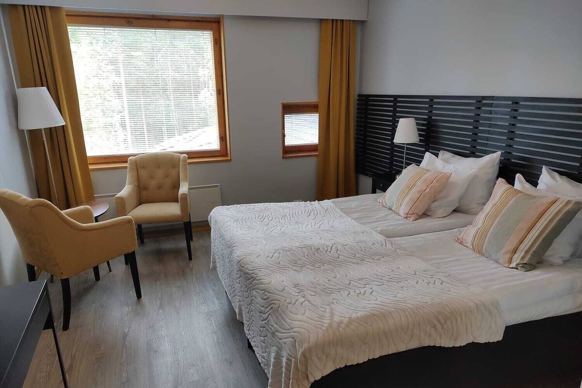 Habitación de hotel con una cama grande, dos mesitas de noche, una silla, un ventanal con cortinas y un cabecero con lámpara.