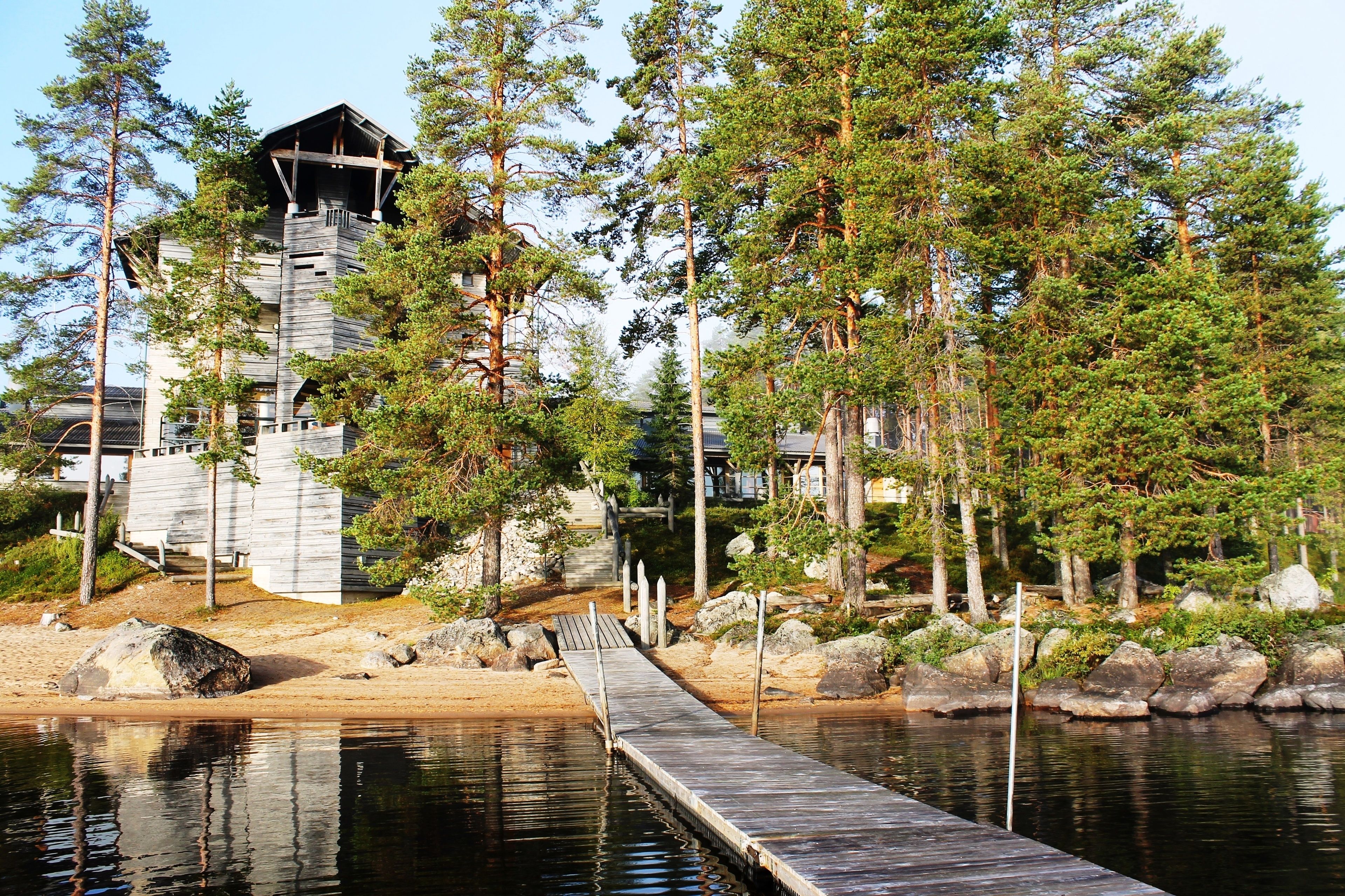 A wooden cabin with a dock extending into a calm lake, surrounded by dense pine trees.