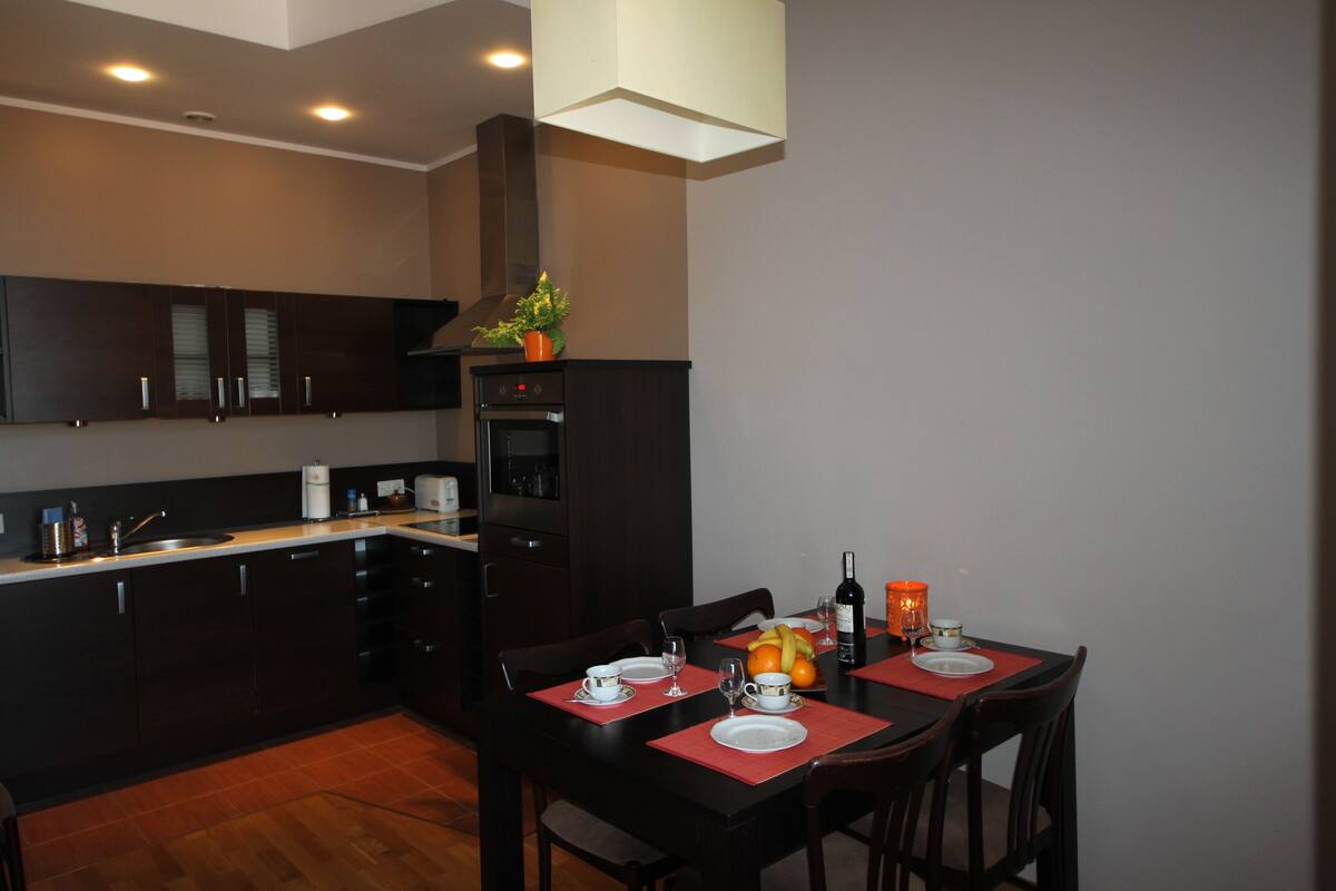 A modern kitchen with dark cabinetry, a central island, and a dining area with red placemats and chairs.