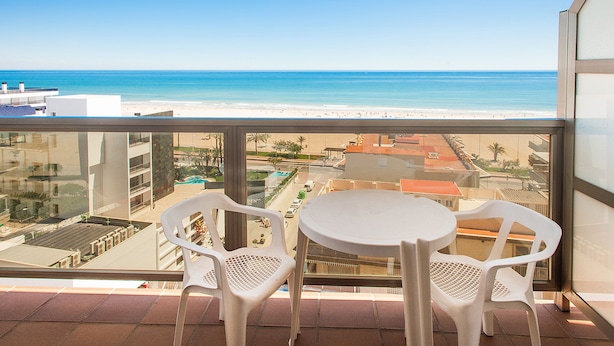 A balcony with two white plastic chairs and a small round table, overlooking a beach with buildings in the background. - Hotel RH Gijón By Hoteles RH (Gandia)