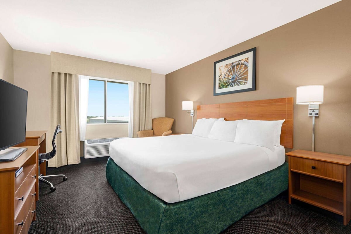 A hotel room with a large bed, a desk with a TV, a chair, a window with curtains, and a wall-mounted lamp.