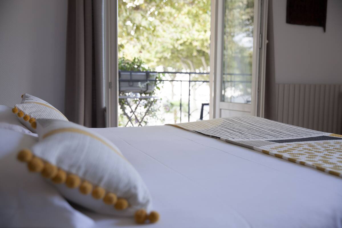 Una cama bien hecha con ropa de cama blanca y detalles amarillos, una ventana con vistas a follaje exterior y un radiador en la pared.