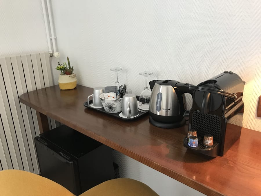 Uma cozinha compacta com cafeteira, chaleira, bandeja com xícaras e copo, e uma pequena planta em vaso.