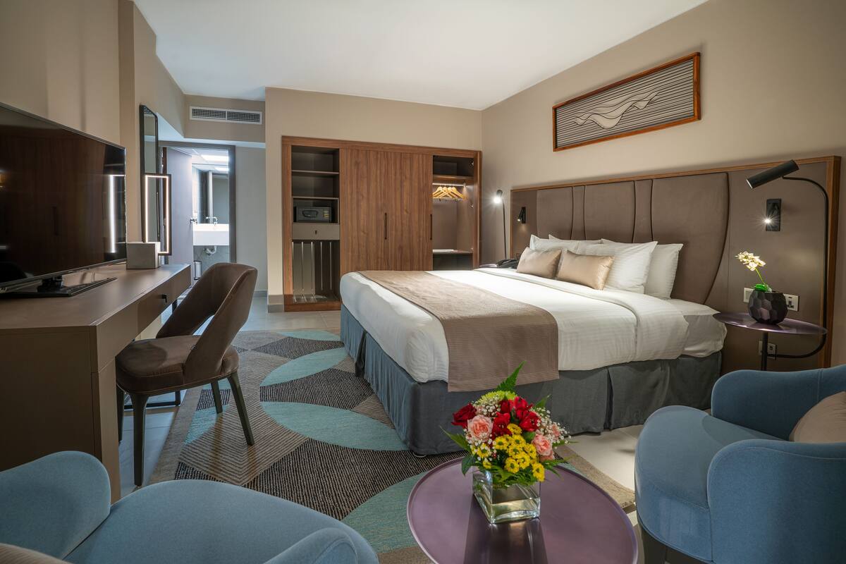 Ein modernes Hotelzimmer mit einem großen Bett, einem Schreibtisch mit Laptop, einem Sessel, einem kleinen Tisch mit Blumenvase und einem begehbaren Kleiderschrank.