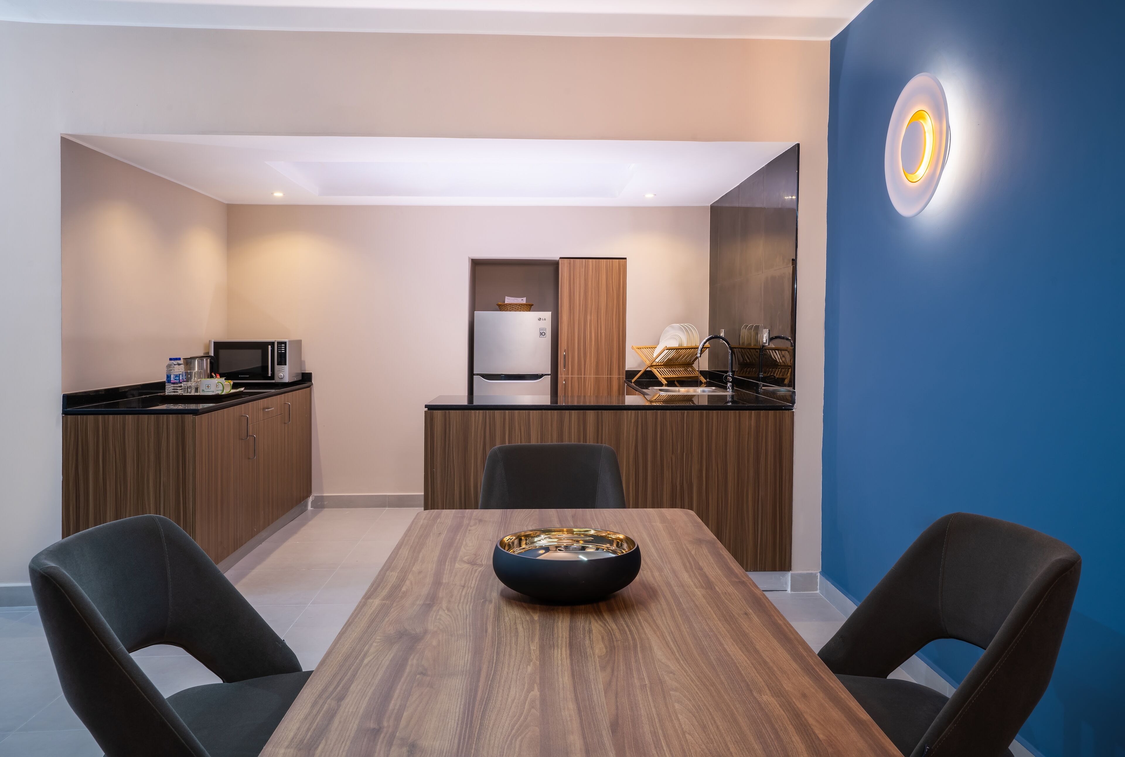 A modern kitchen with wooden cabinets, a microwave, and a refrigerator. There is a dining table with chairs and a decorative bowl in the center.
