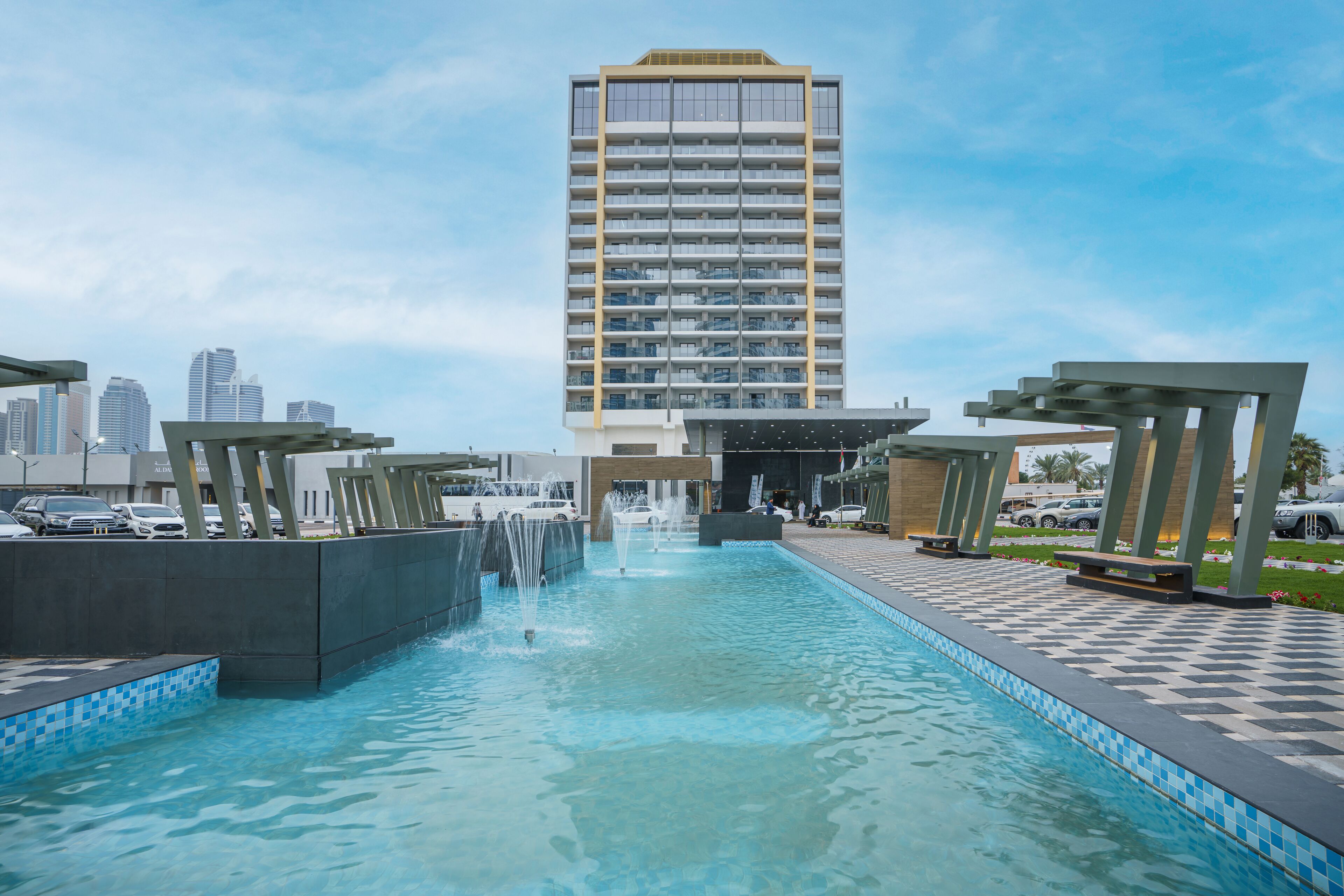 A hotel pool area with a fountain, a checkered walkway, and a tall building in the background.