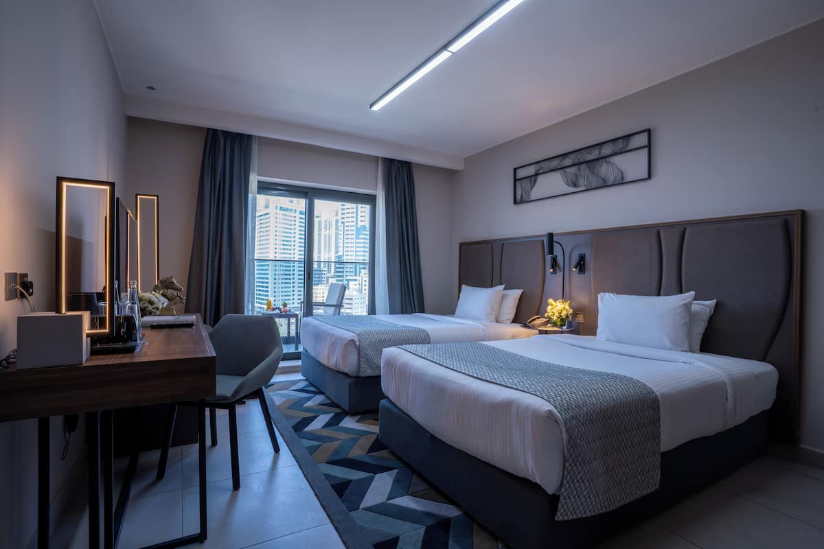 Ein Hotelzimmer mit zwei Betten, einem Schreibtisch mit Computer, einem Stuhl und Blick auf die Stadt durch große Fenster.