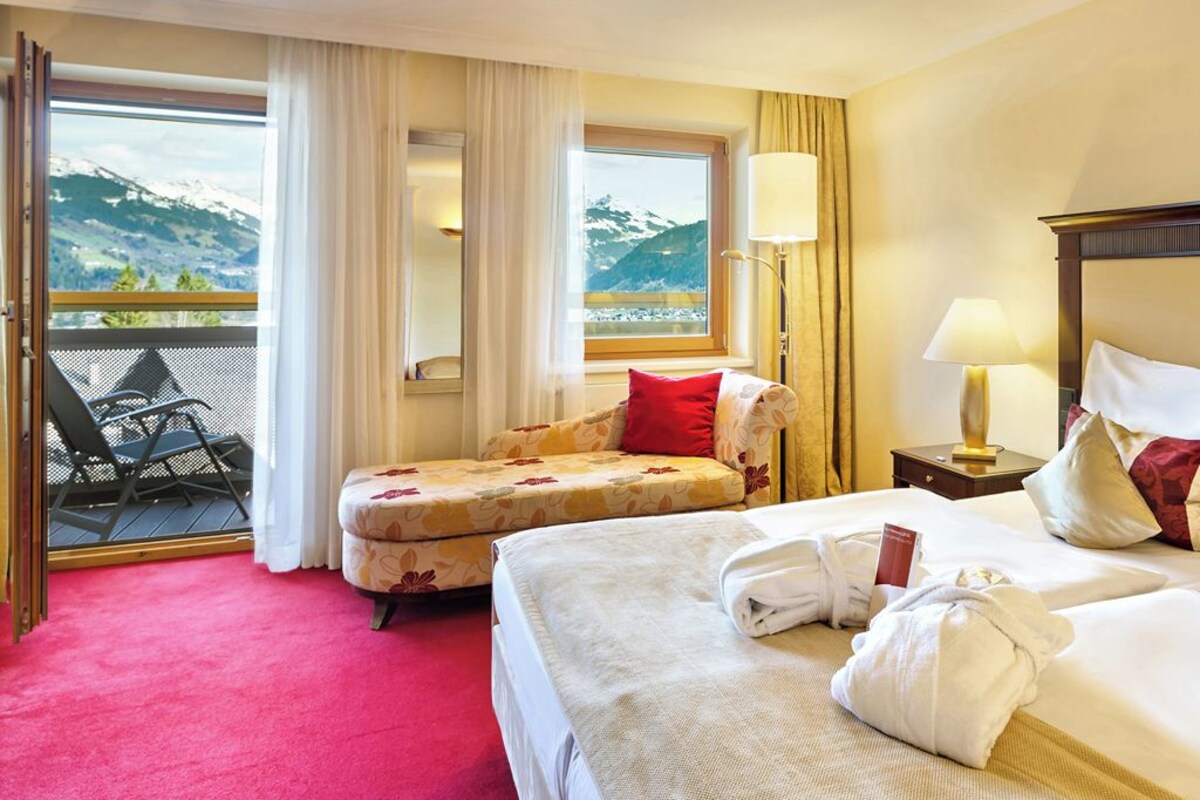 Habitación de hotel con cama, sofá, balcón con sillas y vista a las montañas.