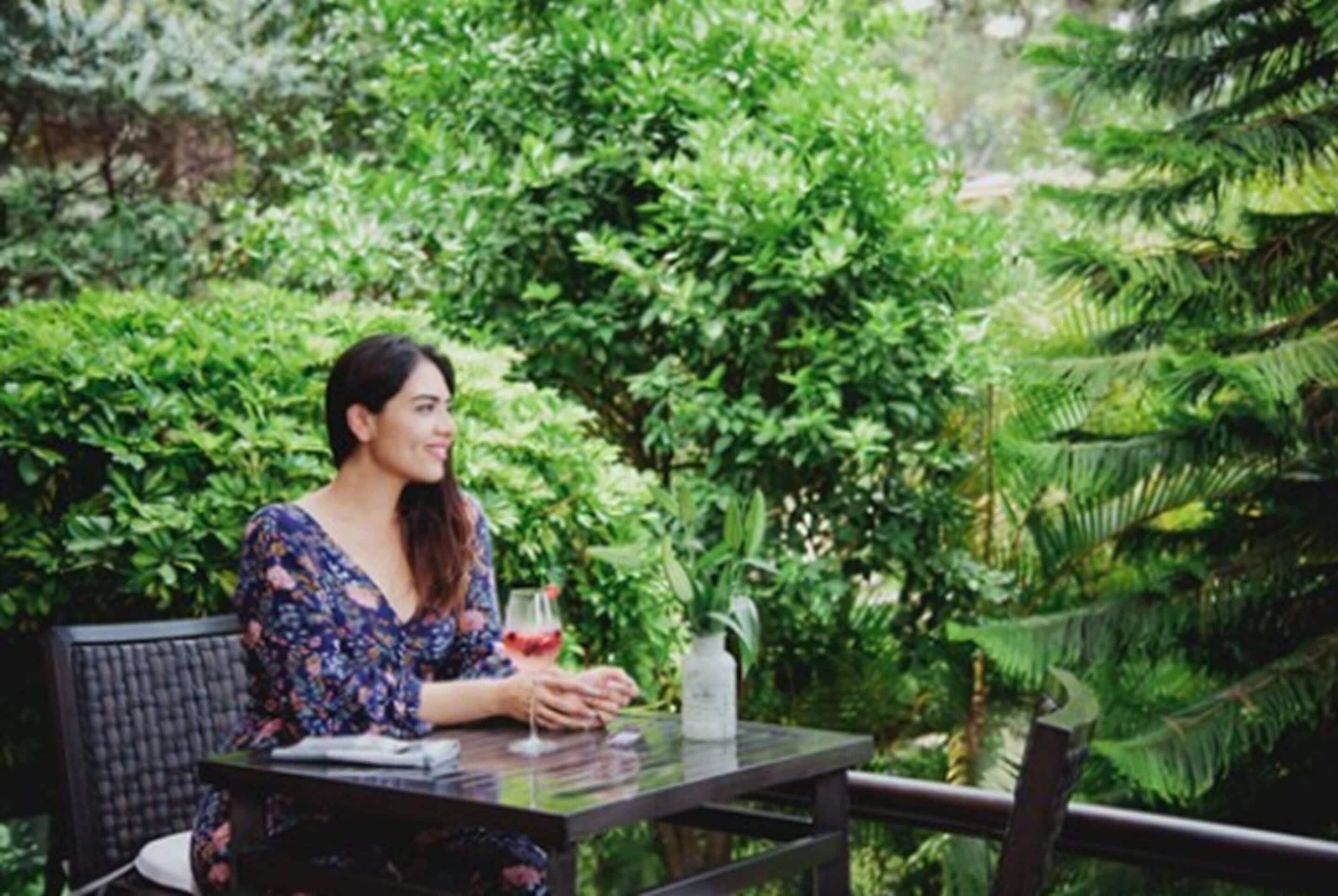Una persona sentada en una mesa al aire libre con una bebida en un vaso, rodeada de abundante vegetación.