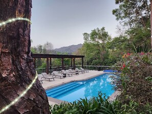 Una piscina con sillones de descanso y una pérgola, rodeada de árboles y colinas.