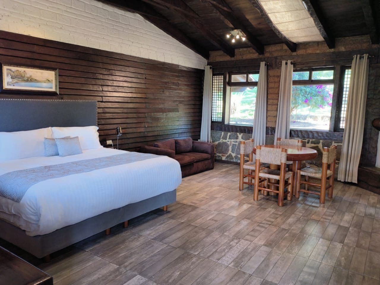 Un dormitorio con una cama grande, cabecera de madera, sofá marrón, mesa de comedor con sillas y pared de piedra.