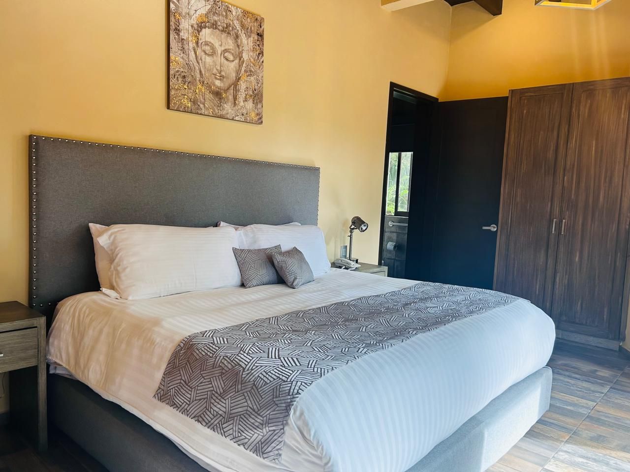 Una cama bien tendida con una colcha estampada y almohadas, una mesita de noche con una lámpara y una puerta a la derecha.