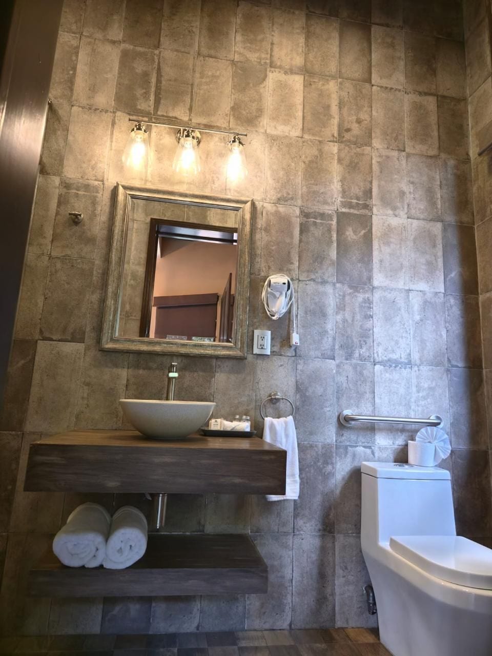 A bathroom with a stone wall, a sink with a vessel basin, a mirror, a towel rack, and a toilet.