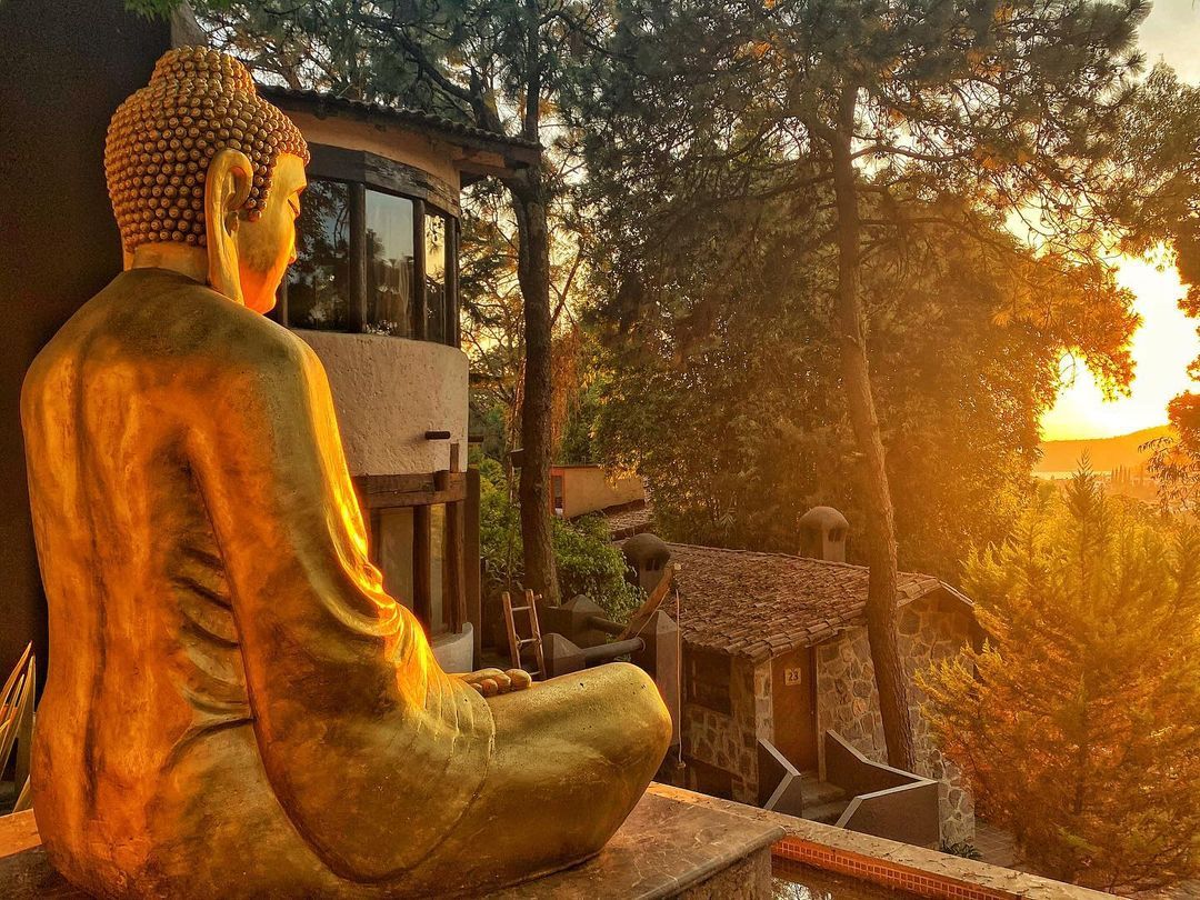 Estátua de Buda em posição sentada, com vista para o pôr do sol e árvores ao fundo.