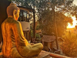 Una estatua de Buda en posición sentada, con vista a una puesta de sol y árboles al fondo.
