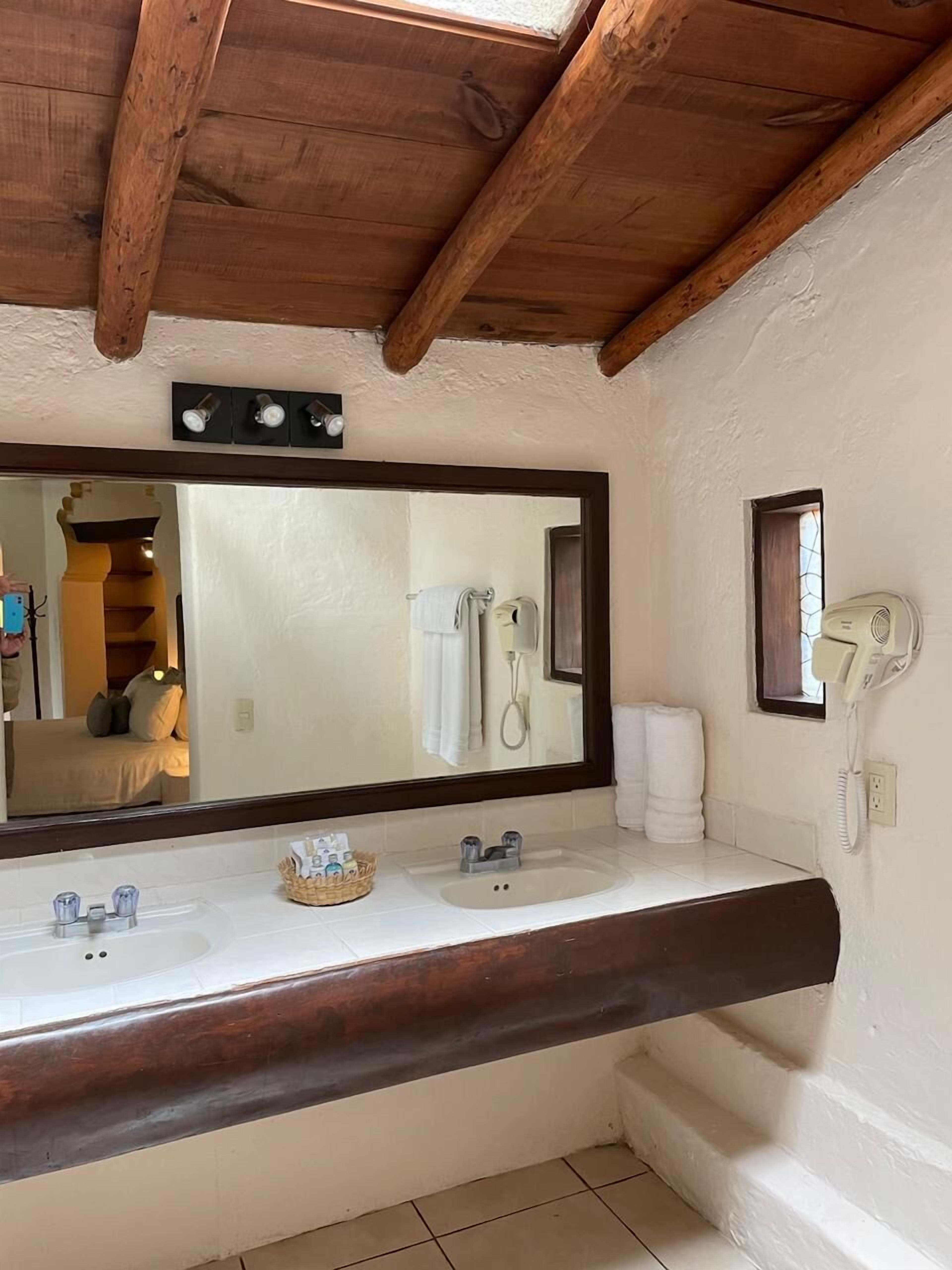 A bathroom with a wooden ceiling, a large mirror, a sink, and a hairdryer on the wall.