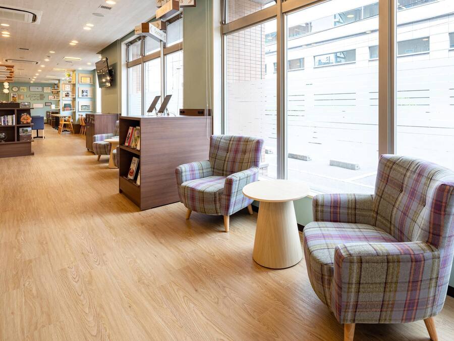 Un café en una biblioteca con sillas a cuadros, una estantería de madera y grandes ventanales.