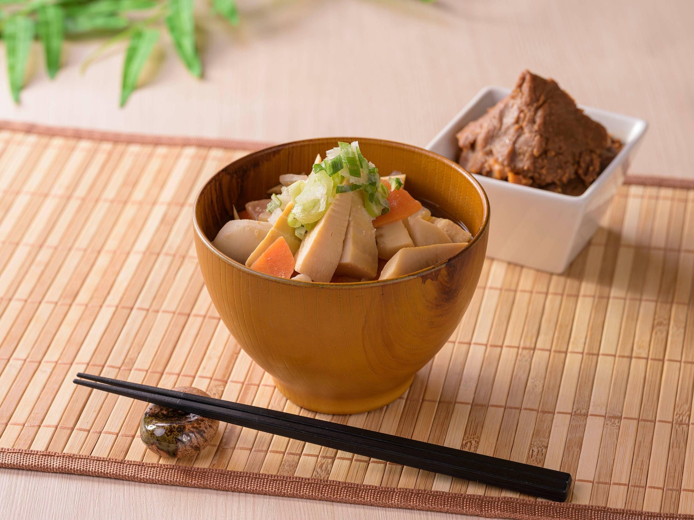Un tazón de tofu y verduras sobre un tapete de bambú, con palillos y un recipiente pequeño de pasta de miso.