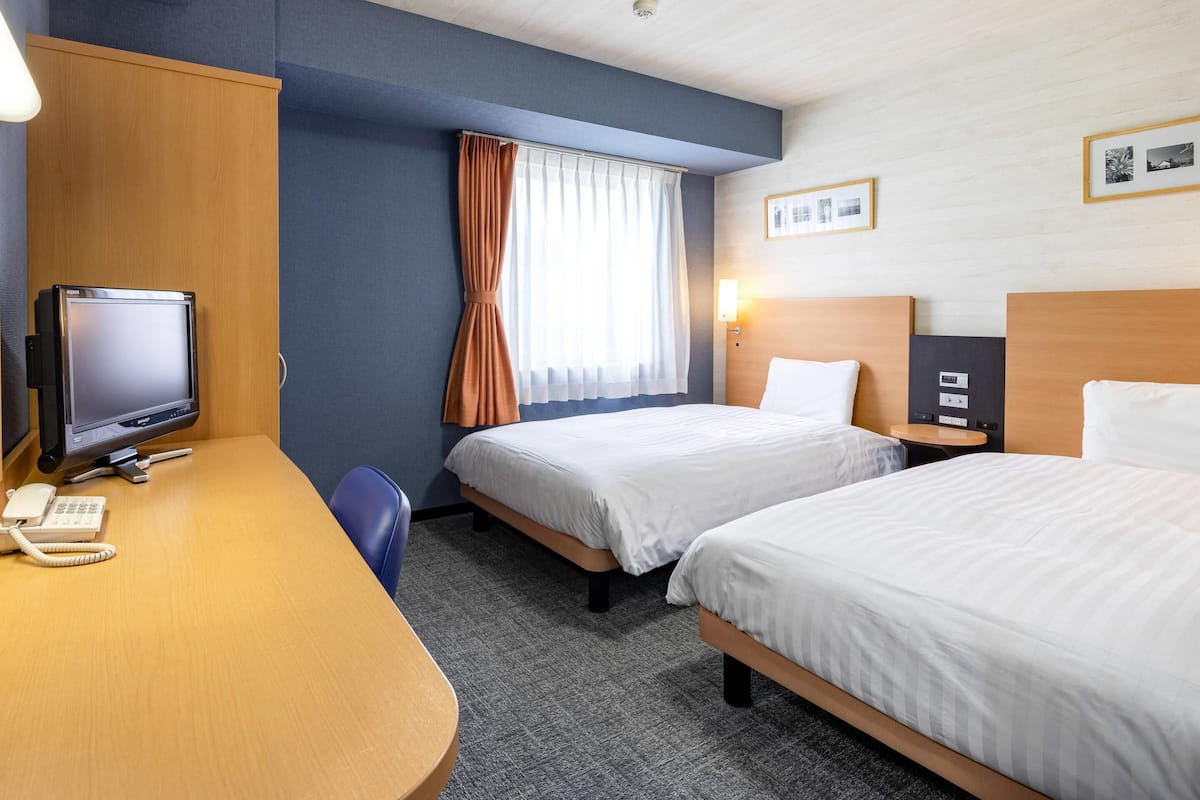 Ein Hotelzimmer mit zwei Betten, einem Schreibtisch mit Computer und Telefon, einem Fernseher und einem Fenster mit Vorhängen.