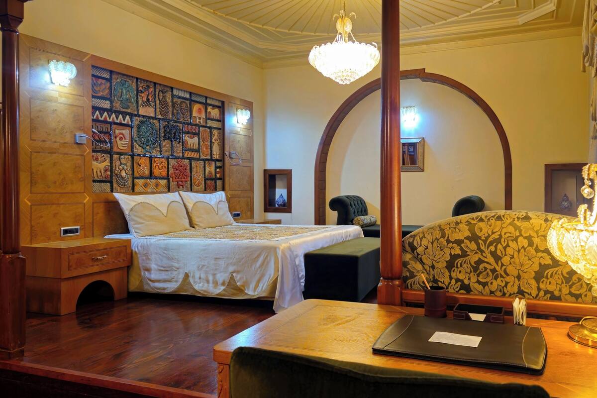 A hotel room with a large bed, a desk with a laptop, a telephone, and a chandelier.