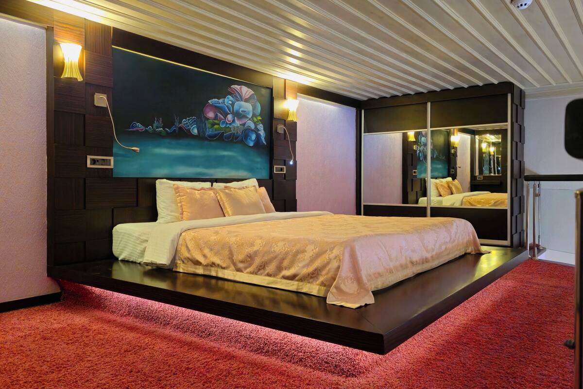 A modern bedroom with a bed centered on a platform, a large flat-screen TV mounted on the wall, and a mirror reflecting the room.