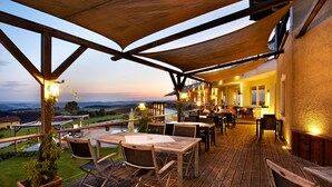 Un patio al aire libre con mesas y sillas de madera, vistas al campo y un edificio con iluminación cálida.