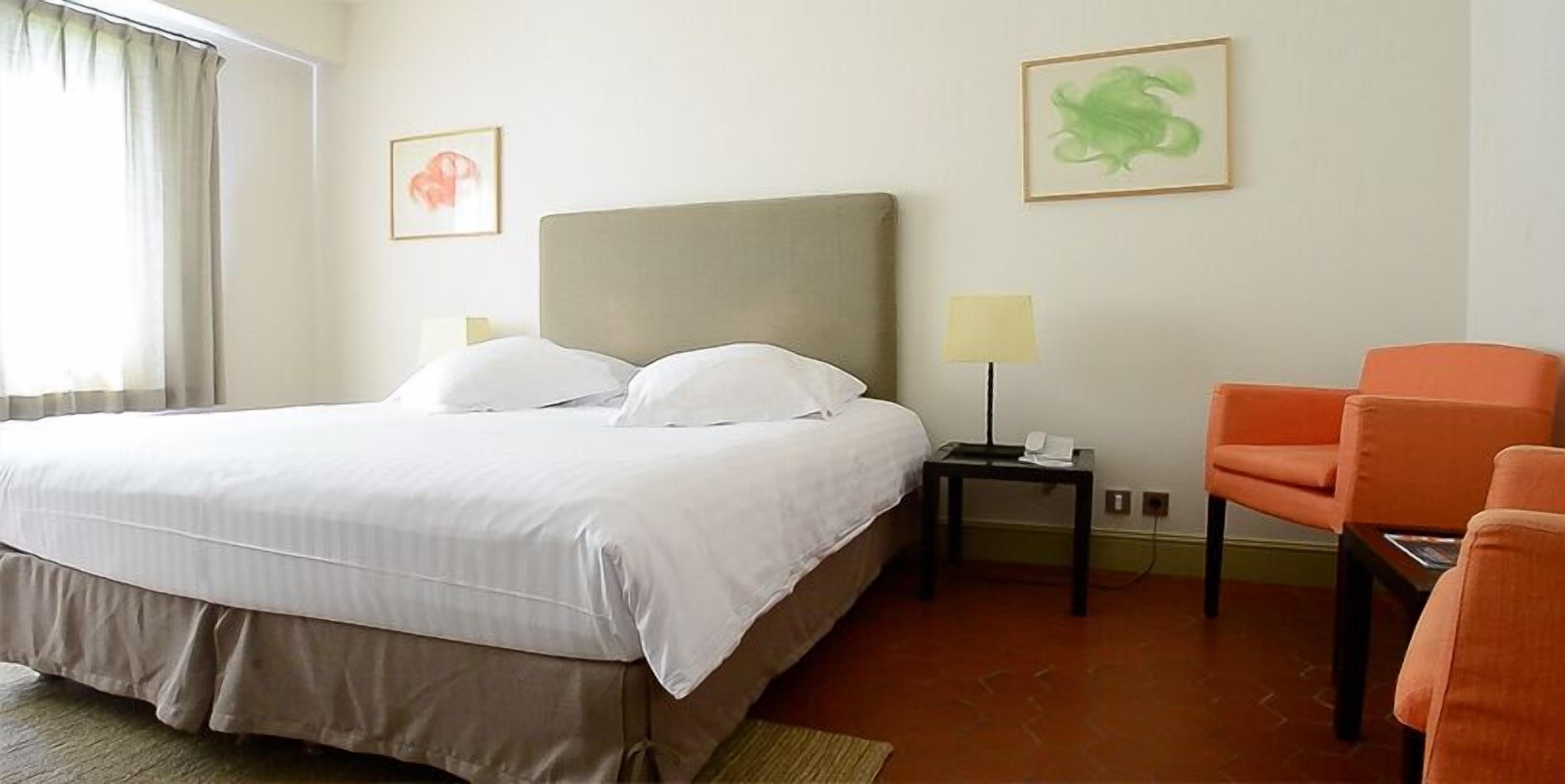 Una habitación de hotel con cama, una silla, una mesita de noche y dos lámparas de pared.