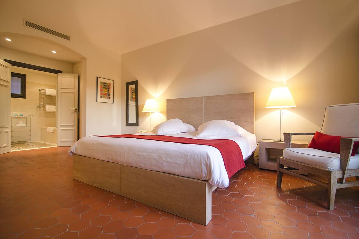 Une chambre d’hôtel avec un grand lit, deux tables de chevet avec des lampes, une chaise en bois et un espace douche.