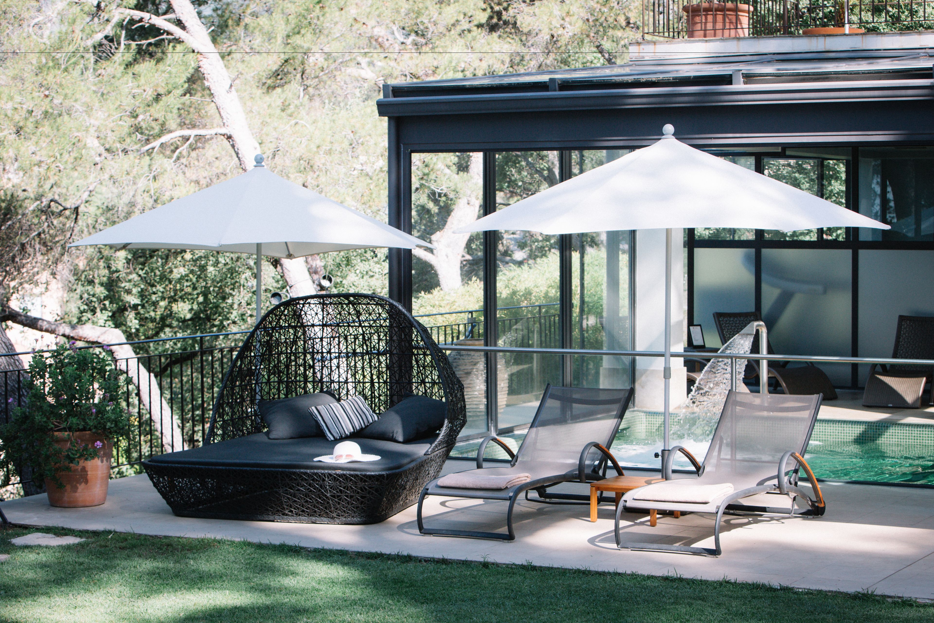 A modern outdoor seating area with a wicker chair and lounge chairs, two umbrellas, and a pool in the background.