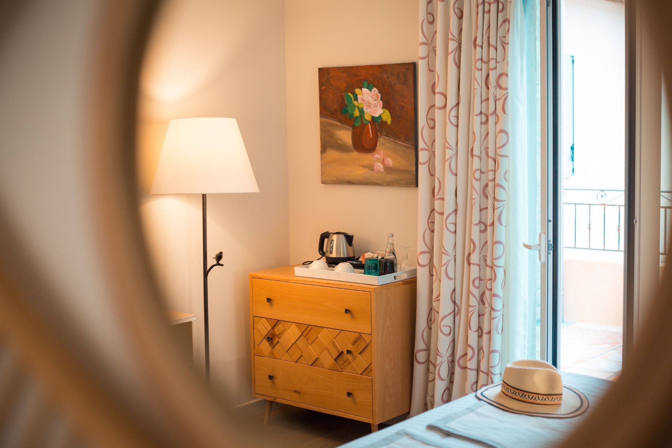 Une chambre d’hôtel avec une table de chevet en bois, une lampe, un tableau de fleurs, une fenêtre avec des rideaux et un chapeau sur le lit.