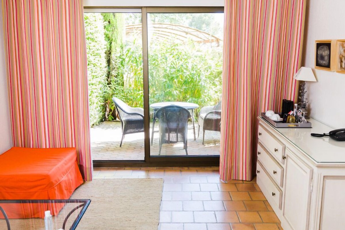A room with a bed, a desk, and a sliding glass door leading to an outdoor area with chairs and greenery.