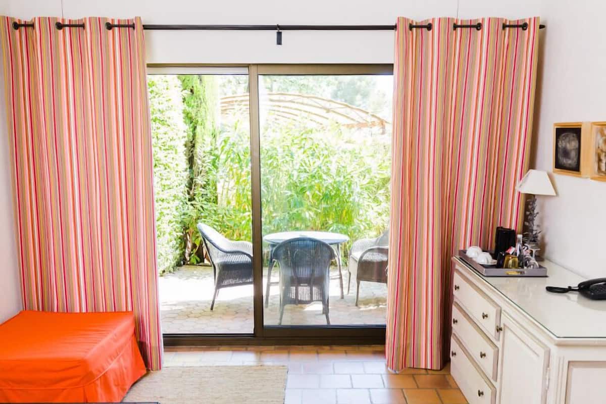 Une chambre comprenant un lit, un bureau et une porte-fenêtre coulissante donnant sur un espace extérieur aménagé avec des chaises et de la verdure.
