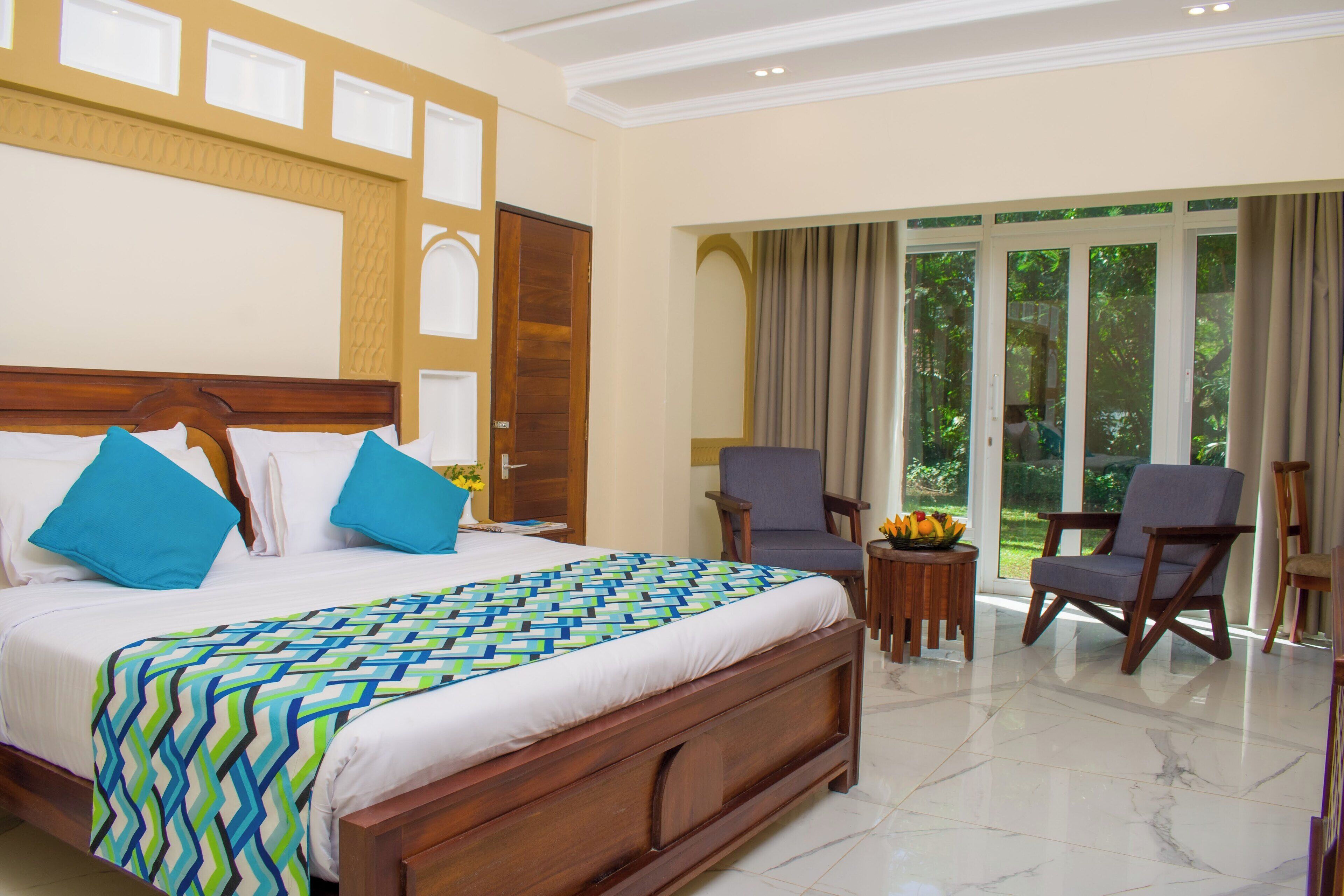 A hotel room with a large bed, two armchairs, a small table with fruit, and a view of greenery through the windows.