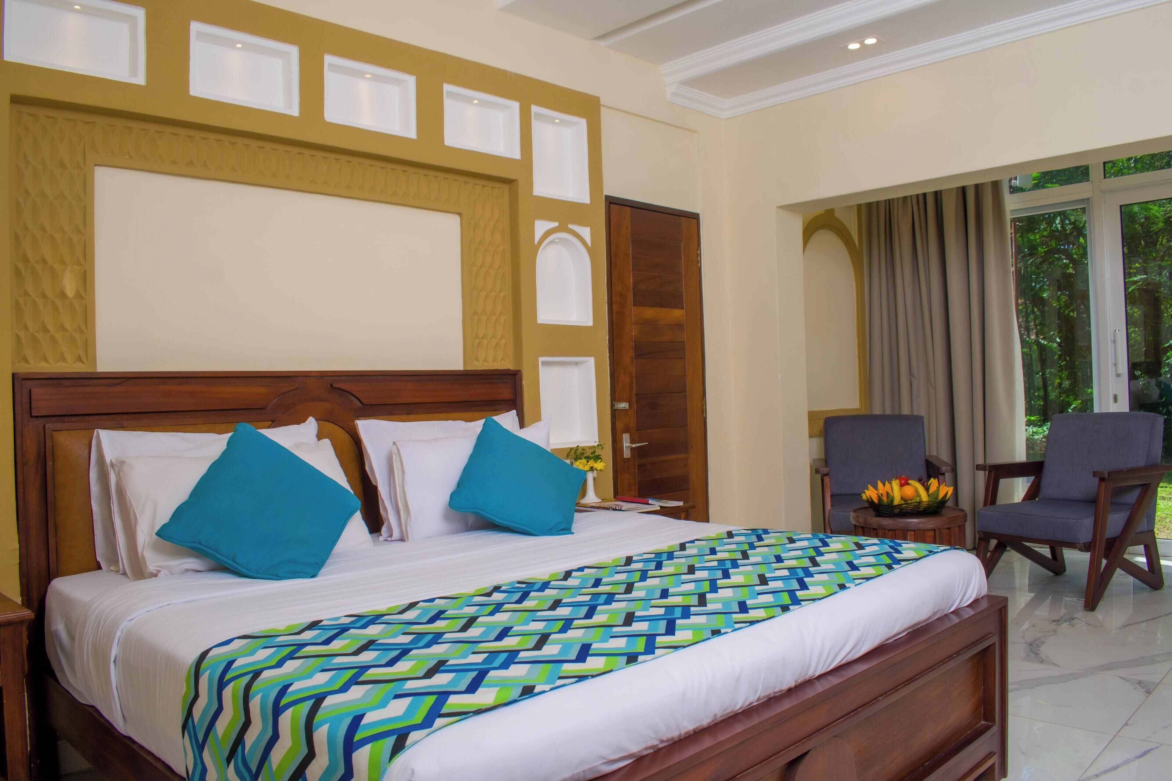 A hotel room with a large bed, two chairs, a small table with a fruit bowl, and a view of greenery through the window.