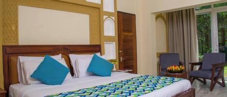 A hotel room with a large bed, two chairs, a small table with a fruit bowl, and a view of greenery through the window.
