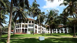 A white building with multiple balconies and arched windows, surrounded by palm trees and a well-maintained lawn. - Cocoa Luxury Resort, Nyali (Mombasa)