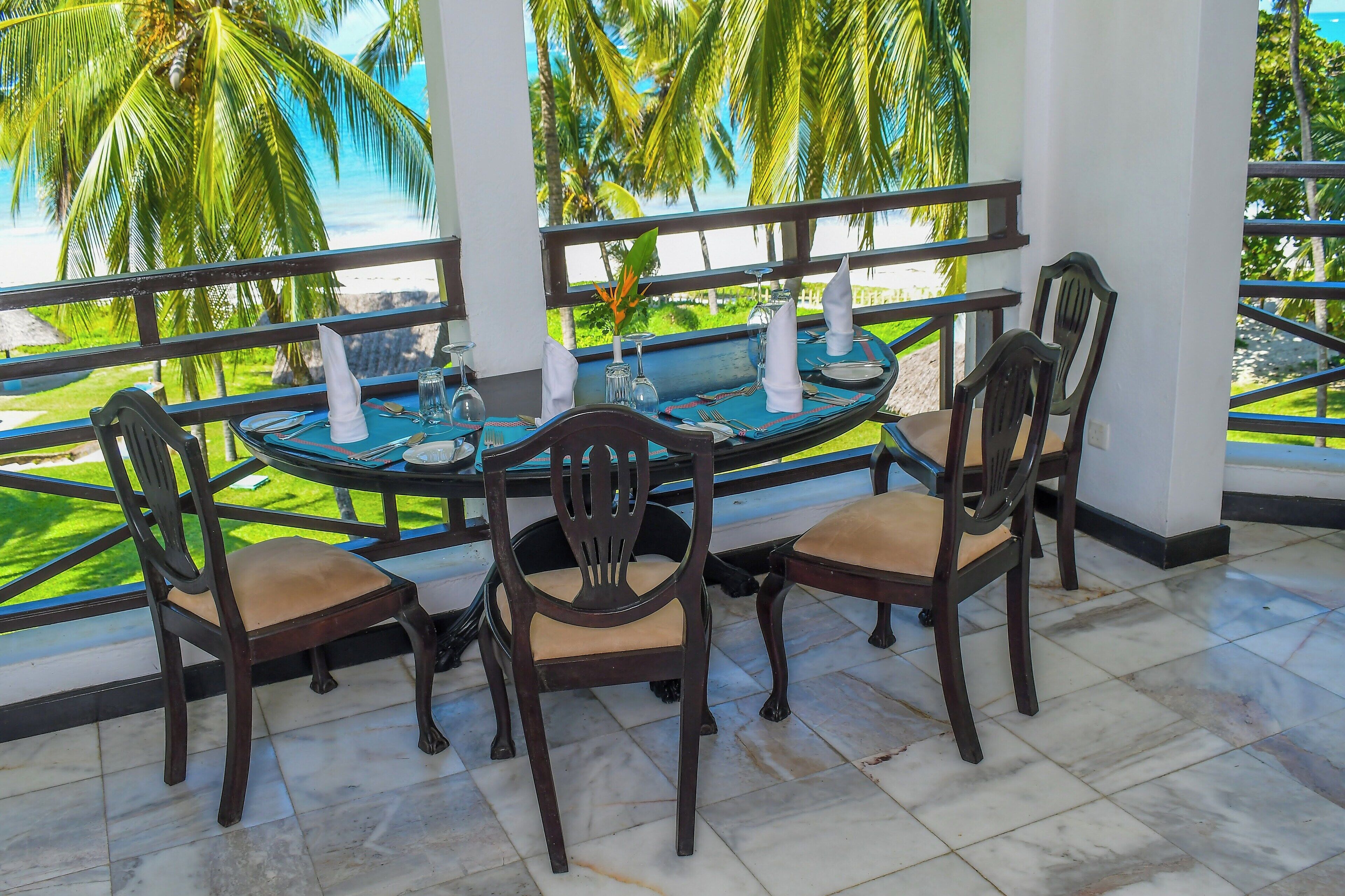 A dining table with four chairs, set with white napkins and glassware, overlooking a tropical landscape with palm trees and a beach.